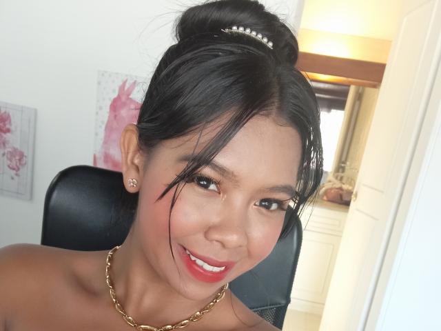 AmiAkira a les cheveux noirs attachés en chignon, avec une pièce de tête en perle, porte une chaîne en or, des boucles d'oreilles en diamant et du rouge à lèvres, elle sourit avec les joues légèrement rouges, assise sur une chaise noire..