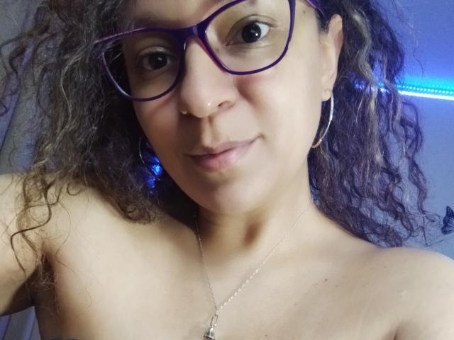 Angelonix with curly brown hair, wearing purple glasses and hoop earrings, has a light brown skin tone, small nose, and is topless, showing bare shoulders and a necklace.