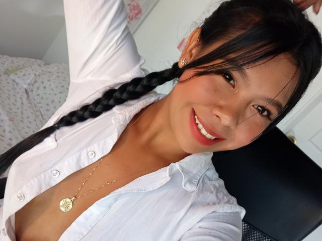 AmiAkira, sorridente, con lunghe trecce nere, indossa una camicia bianca aperta, che lascia intravedere il décolleté, una collana d'oro e orecchini, ed è seduta su una sedia, guardando la fotocamera..