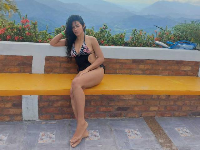OdedySexy, met krullend zwart haar, draagt een zwart en roze bikini met bloemenprint en zit op een gele bakstenen bank. Haar billen en borsten zijn zichtbaar, haar benen zijn gekruist en ze staat zonder schoenen op een betegelde vloer..