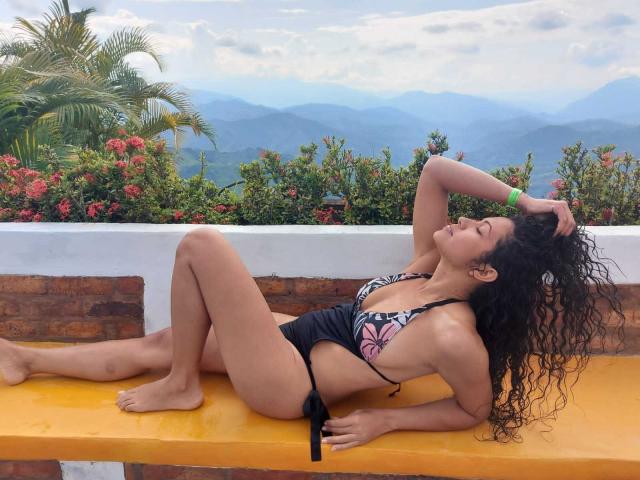 OdedySexy with curly black hair, lying on a yellow bench, wearing a black bikini with pink butterfly, her tits visible, arm raised, legs slightly apart, mountain view background.