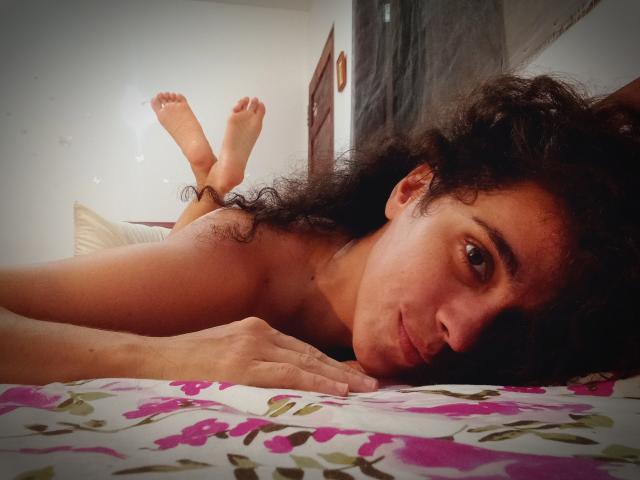 AmyFlores, naakt, ligt op een bed met bloemenprint lakens, heeft krullend zwart haar, bruine huid en een klein achterwerk, haar voeten omhoog, en kijkt met een lichte glimlach naar de camera..