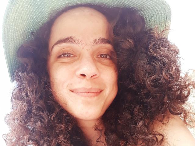 AmyFlores tiene el pelo castaño rizado, ojos verdes y una sonrisa sutil, y lleva un sombrero verde. Su rostro con pecas y su expresión relajada son visibles en la foto, que es brillante y nítida..