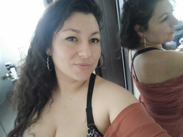 SakotoDika with wavy brown hair, small hoop earrings, and a pink off-shoulder top, showing cleavage and a black bra strap, reflected in a mirror.