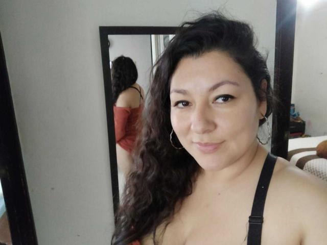 SakotoDika with long, wavy black hair, wearing a black strap top, showing her tits in a mirror reflection, small hoop earrings, and a subtle smile.