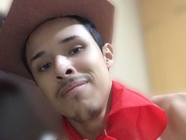Hercolezz tiene un tono de piel marrón claro, cabello negro corto, un sombrero marrón y una camisa roja. Está sonriendo ligeramente y tiene un pequeño bigote, mirando directamente a la cámara..