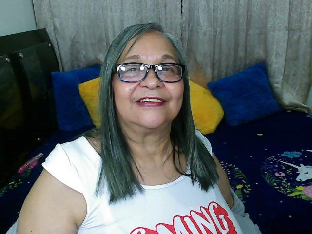 RosarioTriana with gray hair, glasses, white shirt, red text, smiling, medium tits, blue eyes, on dark blue bed with yellow and blue pillows, inside a room.