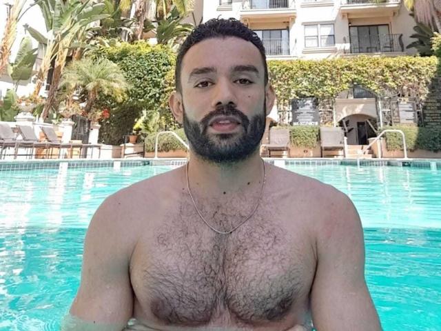 SionActif, shirtless with a hairy chest, stands in a blue pool, looking at the camera, has a beard, and a necklace, with palm trees in the background.