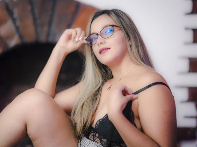 AbrilClark with glasses, long blonde hair, black lace bra, holding strap, slightly smiling, right hand on glasses, left hand on chest, showing her tits, sitting, white nails.