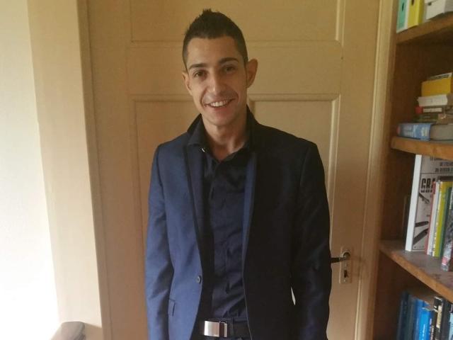 SebSexy with short dark hair, smiling, wearing a navy blue suit over a black shirt, standing in front of a wooden door, right arm behind his back.