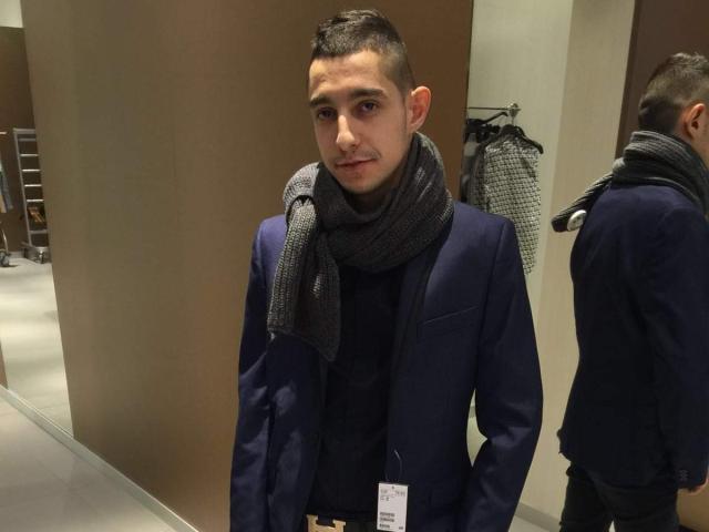 SebSexy with short dark hair, wearing a dark blue suit, black shirt, and gray scarf, standing in a store, reflection in mirror, name tag on suit.