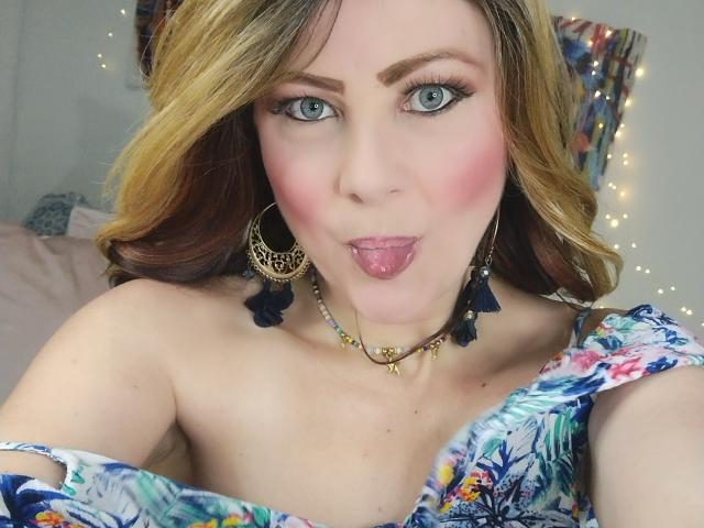 SaraSupreme met blond haar, blauwe ogen, roze blush, tong uitgestoken, gekleed in een bloemenjurk, met gouden oorbellen, een ketting en blauwe oorbellen met een franje, die naar de camera kijkt met een speelse uitdrukking..