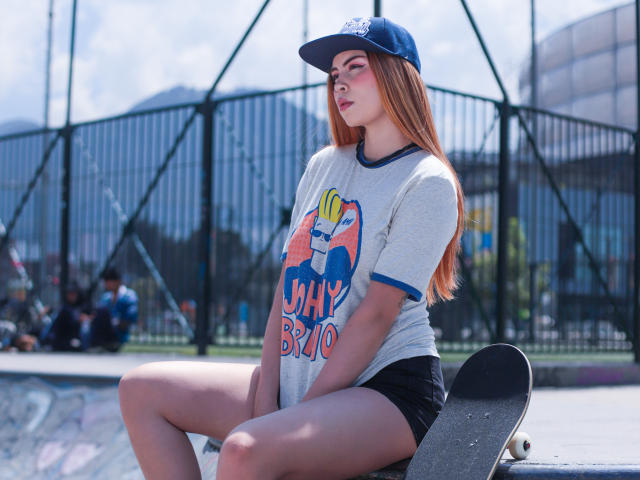 IsaTimeSweet with long red hair, wearing a blue cap and grey "Skay Bro" tee, sitting on a skateboard, black shorts, in a sunny skatepark, legs crossed, looking away.