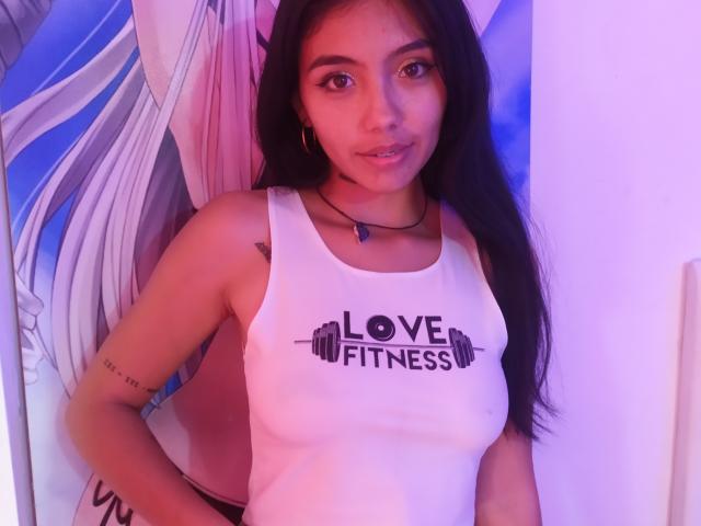 KratosyJuliana, con lunghi capelli neri, indossa una canotta bianca con la scritta "LOVE FITNESS" e ha un seno piccolo, è in piedi in una stanza illuminata di viola e guarda la telecamera con un leggero sorriso..