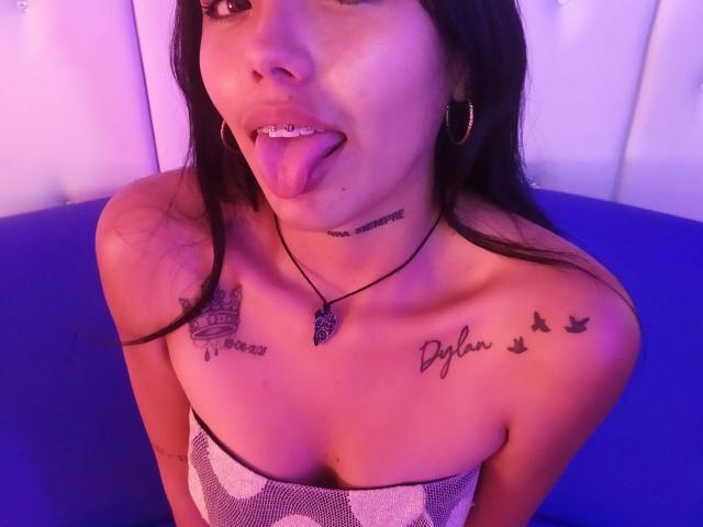 KratosyJuliana, com a língua de fora, mostrando dentes de metal, tem tatuagens no pescoço e nos ombros, usa brincos de argola e está vestindo uma blusa sem alças, sentada em uma luz roxa..