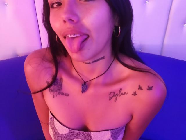KratosyJuliana, Zunge heraus, trägt ein trägerloses Top, zeigt Tattoos auf Brust und Hals, violette Beleuchtung, Creolen und eine schwarze Halskette. Ihre Brüste sind sichtbar..