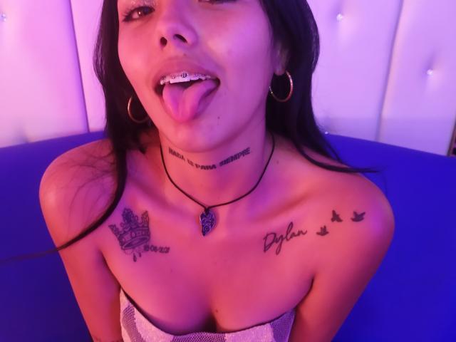 KratosyJuliana with black hair, tongue out, nose ring, hoop earrings, tattoos on neck and shoulders, strapless top, purple-blue lighting, black necklace, and visible cleavage.