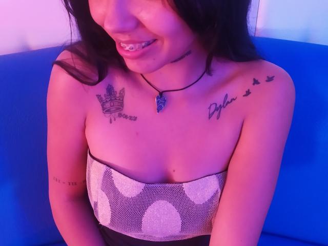 KratosyJuliana, con i capelli scuri, sorridente, indossa un top senza spalline scintillante, mostrando tatuaggi sul petto e sul braccio, una collana blu, seduta su un divano blu, con illuminazione rosa-viola..