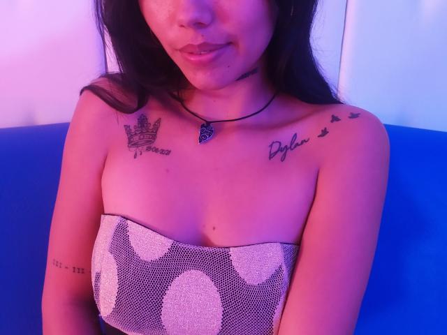 KratosyJuliana has a heart tattoo necklace, black hair, and a strapless top with white polka dots, showing tattoos on her right shoulder and left chest. Her tits are exposed.