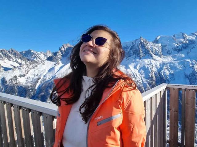 Arleen Hunter, con capelli castano ondulati, occhiali da sole blu, giacca arancione North Face, camicia bianca, sorridente, in piedi su una ringhiera di legno, con cime di montagne innevate sullo sfondo, cielo blu limpido, giornata di sole..