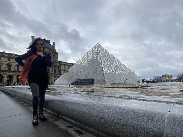 Arleen Hunter, com cabelo escuro, blusa preta e calças de ginástica, segurando um lenço vermelho, em frente à Pirâmide do Louvre, céu nublado, usando botas marrons, calçada cinza..