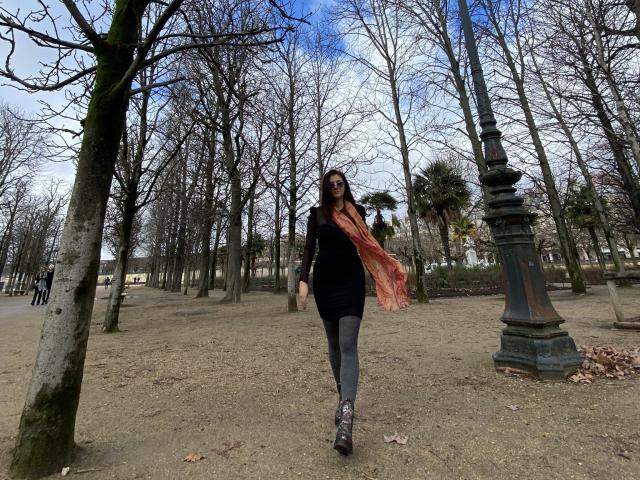 ArleenHunter with long dark hair, black dress, grey leggings, and pink scarf, walking in a park, holding a bag, tree on left, old streetlamp on right, blue sky.