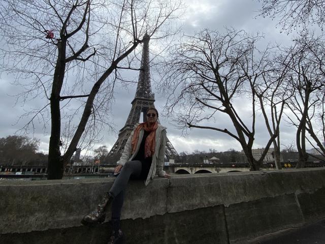 ArleenHunter with tits covered by a pink scarf, sitting on a stone wall, wearing a gray jacket and blue jeans, in front of the Eiffel Tower, cloudy sky.