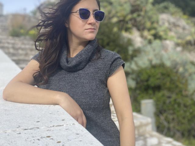 ArleenHunter with wavy brown hair, wearing a gray cowl-neck sweater, blue aviator sunglasses, leaning on a concrete wall, looking slightly to the side with a calm expression.