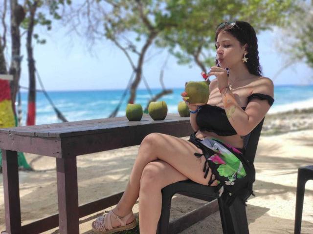 ValeryScot with tan skin, black bikini top, and colorful skirt, sipping from a green coconut, sitting on a black chair, beach table in front, sandy shore, ocean in background.