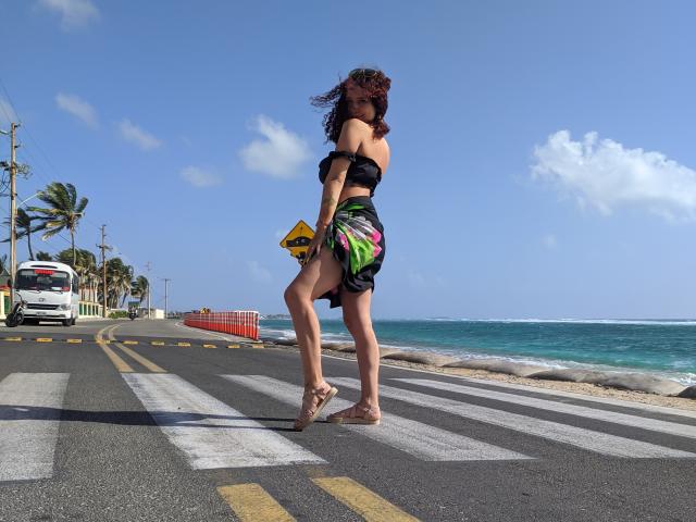 ValeryScot with curly brown hair, black off-shoulder top, and colorful green-black skirt, walking barefoot on a road, holding a yellow sign, ocean and palm trees in background.