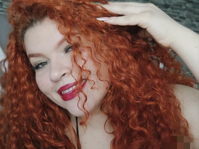 Melida, eine Frau mit lockigem, rotem Haar, lächelt mit rotem Lippenstift, hat grüne Augen und helle Haut. Sie berührt ihren Kopf und zeigt ihr hübsches Gesicht und ihre vollen Brüste..