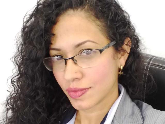OdedySexy ha i capelli neri e ricci, porta gli occhiali e ha il rossetto rosa. Indossa un completo grigio a righe, con orecchini dorati, e ha un'espressione leggermente seria..