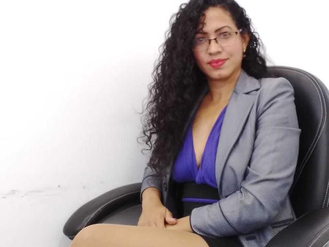 OdedySexy, com cabelos pretos encaracolados e óculos, está sentada em uma cadeira preta, vestindo um blazer cinza riscado e um vestido azul, mostrando seus seios em um decote em V profundo..