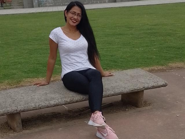 OdedySexy with long black hair, glasses, white polka-dot shirt, black pants, pink Nike sneakers, sitting on a concrete bench, smiling, showing her ass, in a grassy park.