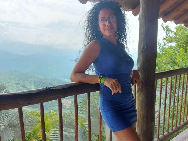 OdedySexy, avec ses cheveux noirs bouclés, ses lunettes et sa robe bleue, se tient contre une rambarde en bois, montrant ses fesses et ses petites seins, et porte un bracelet vert, tout en souriant..