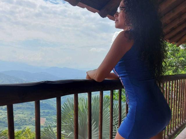 OdedySexy, con cabello negro rizado, vestida con un vestido azul ajustado, apoyada en un balcón de madera, mostrando su trasero, mirando una vista de la montaña, bajo un porche cubierto..