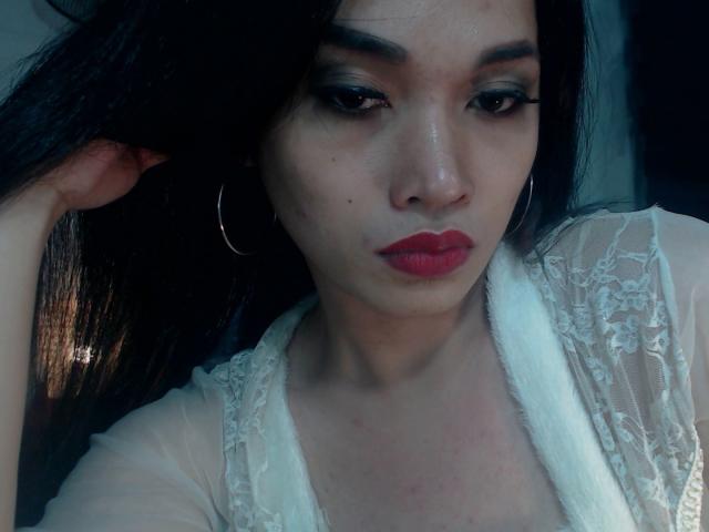 CuteBiggTitsX with dark hair, red lipstick, hoop earrings, and white lace top, looking down with a slight pout, hand in hair, slightly shiny skin, soft lighting.