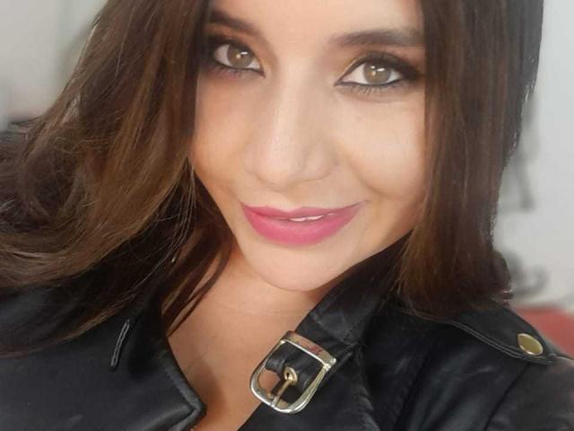 Sharlote Durant, com olhos castanhos, batom rosa e jaqueta de couro preta, sorrindo para a câmera, usando um cinto na jaqueta, tem cabelo castanho de comprimento médio, e parece linda e sexy..