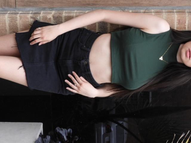 IsaTimeSweet met lang zwart haar, een groene crop top, een zwarte rok en een sterrenketting, staat, toont haar buik, heeft haar hand op haar heup en heeft een subtiele glimlach, met een houten muur op de achtergrond..