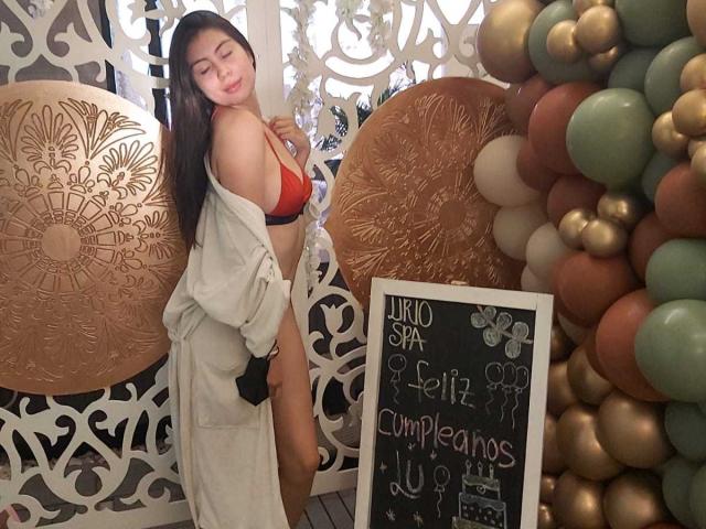 IsaTimeSweet met lang zwart haar, een rode bh en een open gewaad, die haar borsten en billen laat zien, staat in de buurt van een ballon en een decoratieve muur. Ze glimlacht met gesloten ogen..