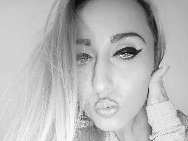 AminaSecrets with pouty lips, half-shut eyes, and thick black eyeliner. She has long, slightly messy hair, and her hand is touching her cheek. Black-and-white photo.