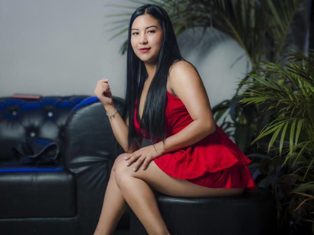 MeganAndHanna, sitting on a black leather couch, wearing a red dress, showing her ass, with long black hair, and a subtle smile, looking at the camera.