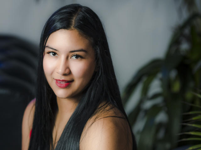 MeganAndHanna with long black hair, dark eyes, and red lipstick, smiling slightly at the camera, wearing a red top, her small tits visible, in a well-lit photo.