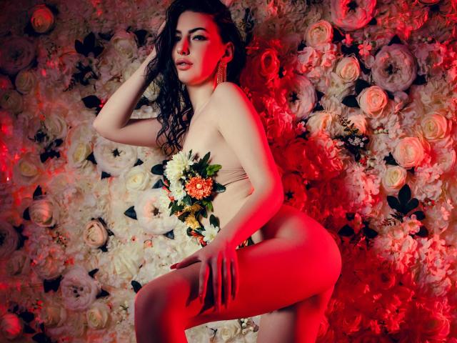 LucyGrace with wavy dark hair, nude, holding a bouquet of flowers, covering her tits with her hand, red light on her ass, against a floral wall.