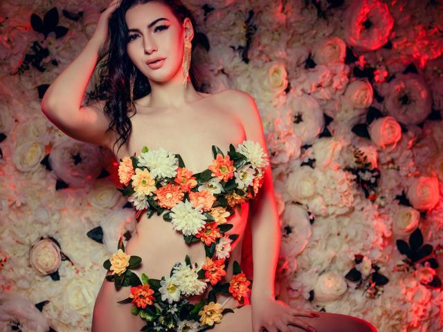 LucyGrace con flores cubriendo sus pechos y vulva, usando adornos de hojas verdes, de pie frente a una pared de rosas blancas, con una mano en su cabello oscuro y ondulado, iluminación roja..