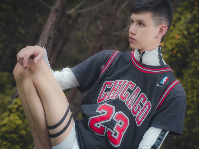 JeanKarloLeone mit kurzen, schwarzen Haaren, der ein Chicago 23 Trikot, ein weißes Langarmshirt und graue Shorts trägt, sitzt mit angezogenen Knien und blickt zur Seite, in einem Wald..