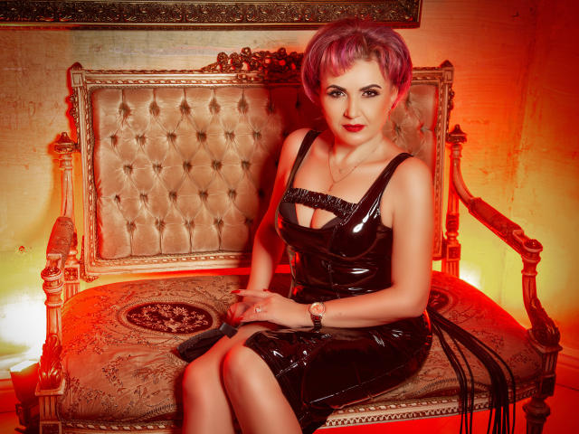 IsabelaConte sits on an ornate, vintage sofa, wearing a shiny black latex dress that shows her tits, with purple hair and red lighting. Her legs are crossed.