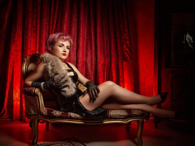 IsabelaConte lounges on a gold chair, legs crossed, wearing black latex, furry vest, gloves, and heels. She has pink hair, red lipstick, and a sexy, confident look.