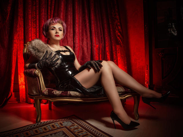 IsabelaConte with pink hair, black latex dress, and gloves, sitting on a chair, legs crossed, wearing black heels, red light, fur trim, and patterned rug. Boobs, ass, and legs visible.