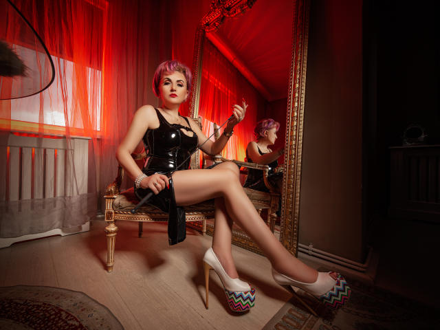 IsabelaConte with pink hair, black latex dress, and white heels, sitting on a chair, holding a whip, reflection in a large mirror, red lighting, legs crossed, looking at camera.